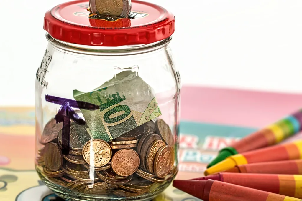 Coins and a banknote filling a glass savings jar, with another coin dropping through the lid