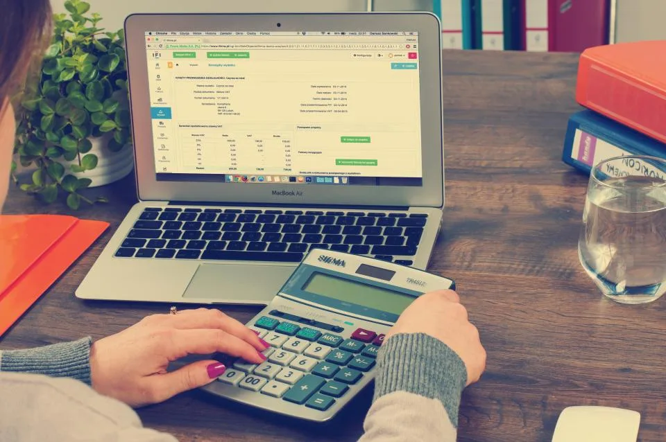 A person working out figures on a desktop calculator beside a laptop showing a VAT invoice